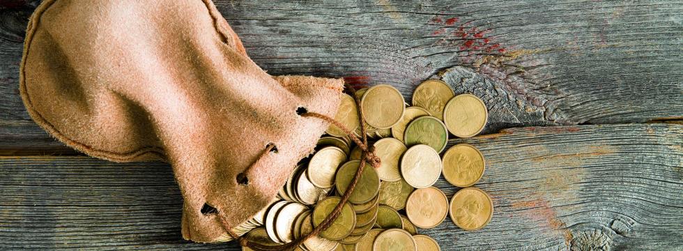 Ein Sack mit Münzen liegt auf einem Untergrund aus Holz und es fallen goldene Münzen heraus