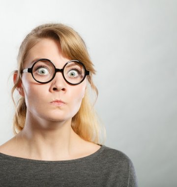 Eine Frau mit blondem Haar, großer runder schwarzer Brille und grauem Oberteil blickt überrascht und mit großen Augen in die Kamera vor einem schlichten Hintergrund.