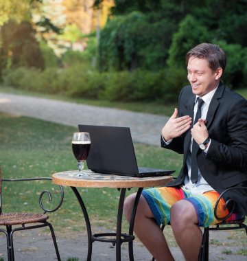 Mann sitzt im freien in Krawatte und Anzug aber mit kurzer Sommerhose in einem Online Meeting mit einem Bier hinter dem Laptop