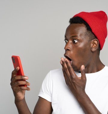 Eine Person, die eine rote Mütze und ein weißes T-Shirt trägt, schaut mit einem überraschten Gesichtsausdruck auf ein Smartphone und hält sich eine Hand vor den Mund.