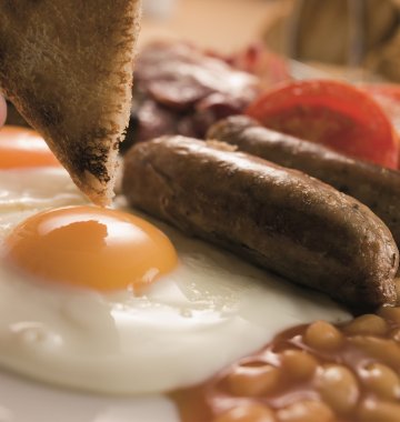 Nahaufnahme eines Frühstückstellers mit Spiegeleiern, Würstchen, Baked Beans, gegrillter Tomate und einer Hand, die ein Stück Toast hält.