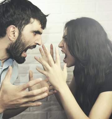 Ein Mann und eine Frau stehen sich vor einer weißen Backsteinmauer gegenüber, beide schreien mit ausdrucksstarker Miene und erhobenen Händen.