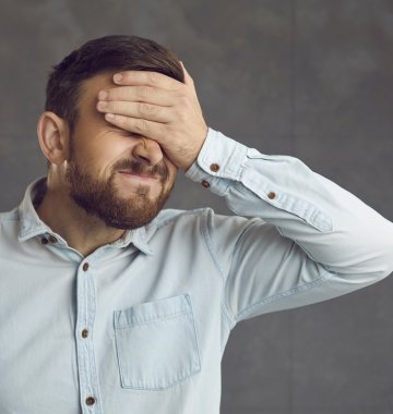 Ein Mann in einem hellblauen Hemd steht vor einem grauen Hintergrund und verdeckt mit der Hand sein Gesicht in einer Geste der Frustration oder Enttäuschung.