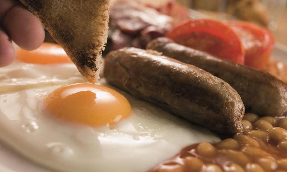 Nahaufnahme eines Frühstückstellers mit Spiegeleiern, Würstchen, Baked Beans, gegrillter Tomate und einer Hand, die ein Stück Toast hält.
