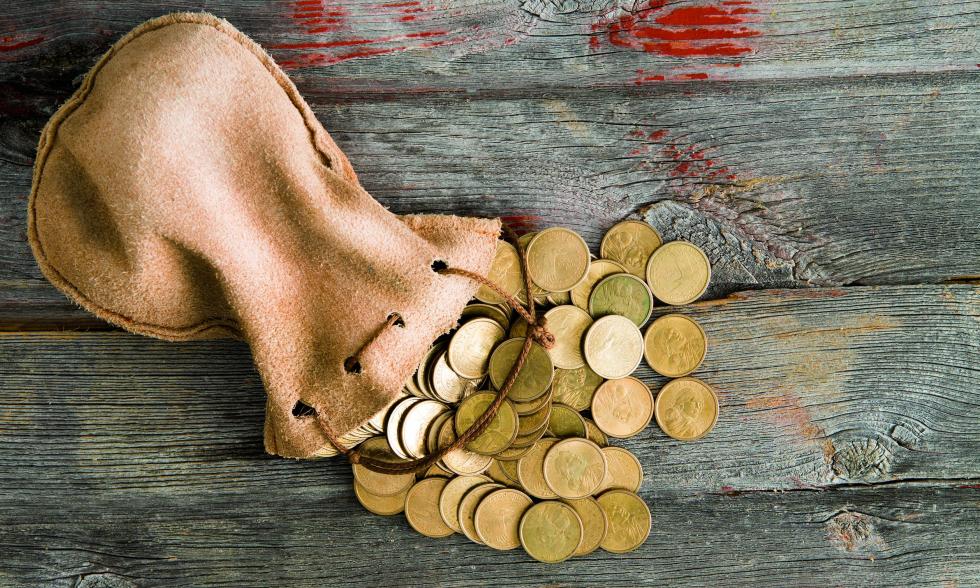 Ein Sack mit Münzen liegt auf einem Untergrund aus Holz und es fallen goldene Münzen heraus