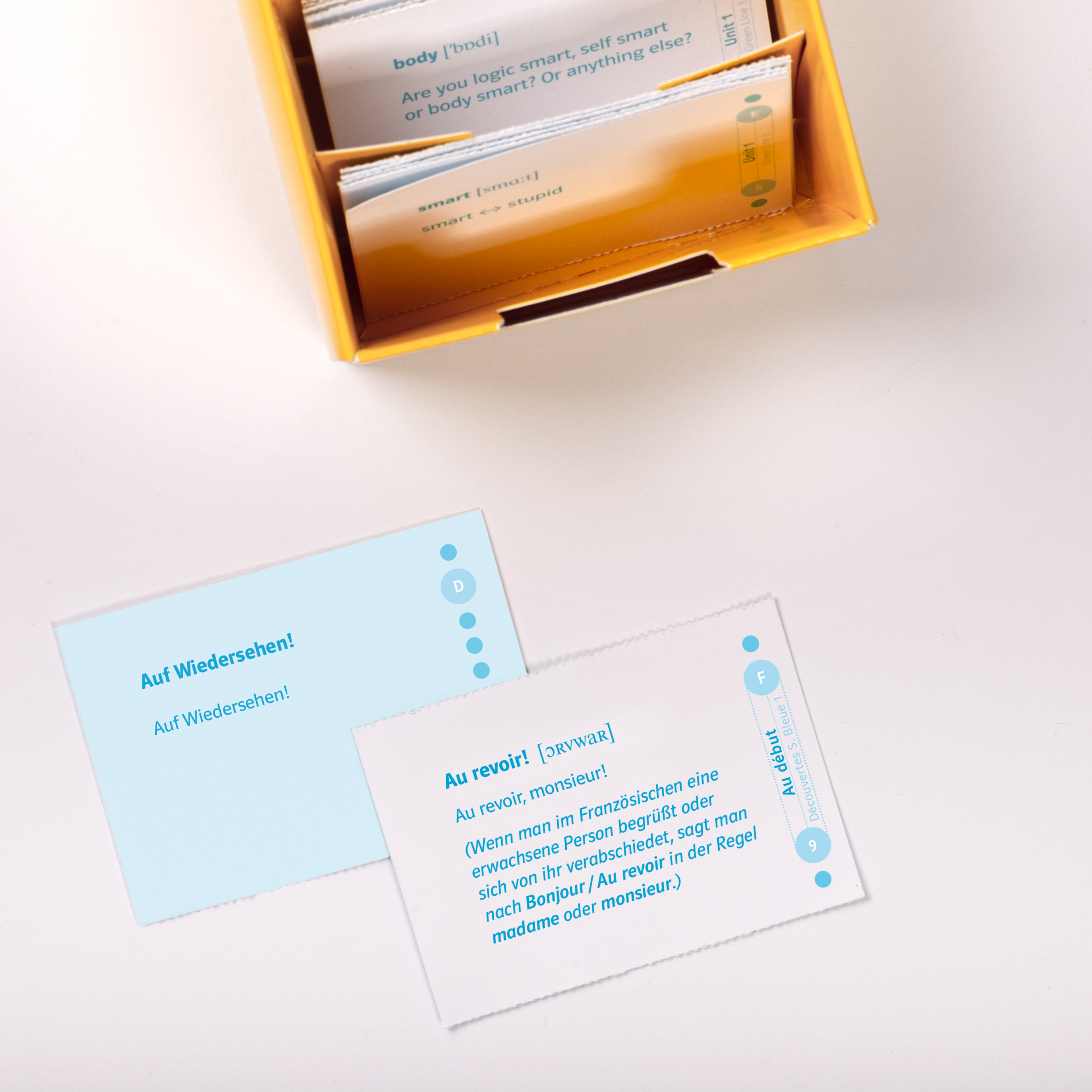 Eine gelbe Schachtel enthält Sprachlernkarten, wobei zwei Karten auf einer weißen Fläche die Sätze Auf Wiedersehen! auf Deutsch und Au revoir! auf Französisch abbilden.